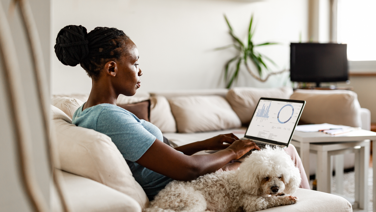 Woman working from home using a laptop on the couch and seated next to a small dog